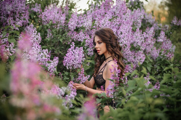 a beautiful girl with makeup and hair styling in underwear stands in a garden with lilacs on a summer evening