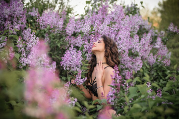 a beautiful girl with makeup and hair styling in underwear stands in a garden with lilacs on a summer evening