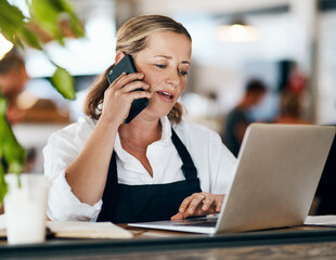 Coffee shop owner on a phone call while working online on her laptop inside a local cafe store....