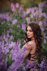 a beautiful girl with makeup and hair styling in underwear stands in a garden with lilacs on a summer evening