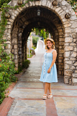 Fototapeta na wymiar Portrait of a pretty young woman in a blue dress and straw hat. Summer vacation.Background of an old historical stone arch building