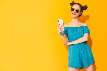 Young beautiful smiling female in trendy summer green dress. Sexy  woman with posing near yellow wall in studio. Positive model having fun. In sunglasses. Looking at smartphone. Using apps