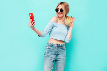 Young beautiful smiling blond female in trendy summer jeans clothes. Sexy carefree woman posing near blue wall in studio. Positive model having fun indoors. Cheerful and happy. taking selfie