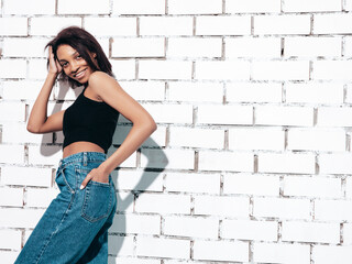 Portrait of young beautiful black woman. Smiling model dressed in summer jeans clothes. Sexy carefree female posing near white brick wall in studio. Tanned and cheerful