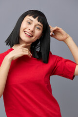 a sweet, beautiful, attractive woman stands on a gray background in a red T-shirt and touches her hair with a gentle hand gesture, smiling pleasantly