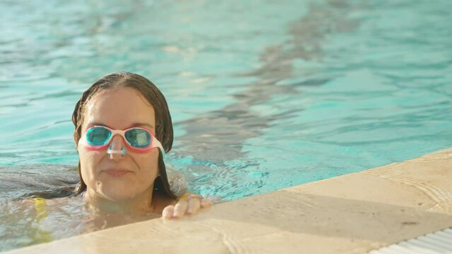 A girl in the pool pops up from under the water, wearing pink glasses and with a clip on her nose, close-up, slow motion