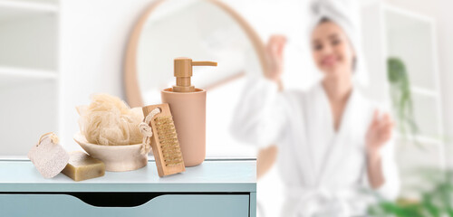Set of bathing supplies on table in bathroom. Young woman in bathrobe and with cosmetic oil - obrazy, fototapety, plakaty