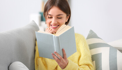 Beautiful young woman reading interesting book on sofa at home