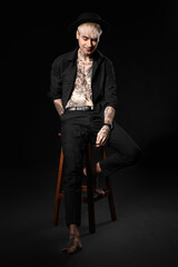 Obraz na płótnie Canvas Young tattooed man sitting on stool against black background