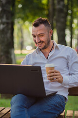 Smiling businessman is working in the park with a laptop, drinking coffee. A young man on a background of green trees, a hot sunny summer day. Warm soft light, close-up.