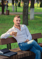 Obraz premium A pensive man sits on a bench, drinks coffee, holds dollars. A young man on a background of green trees, a hot sunny summer day. Warm soft light, close-up.