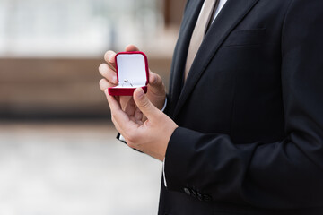 Cropped view of man in elegant suit holding box with engagement ring outdoors.