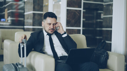 Tilt up of confident attractive businessman talking mobile phone and using laptop computer while sitting on armchair in luxury hotel with luggage suitcase near him. Travel, business and people concept