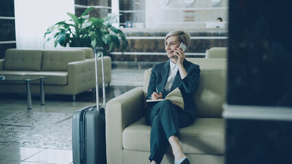 Attractive cheerful businesswoman sitting in armchair in hotel lobby talking on mobile phone and writing in notepad smiling and looking aside. Business, travel and people concept