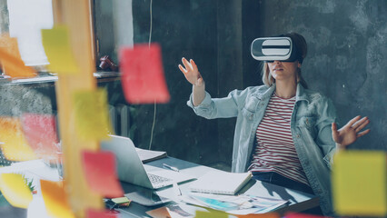 Attractive young woman is sitting at table and wearing virtual reality glasses. She is moving hands and and having VR experience in modern lof office indoors