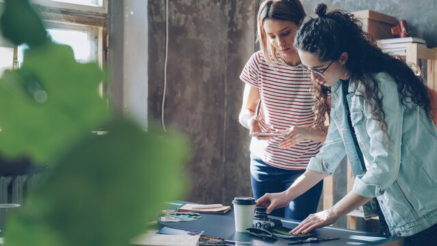 Pretty young women are standing near table, placing colorful pictures on it and shooting flat lay with smartphone. They are sharing ideas about design and smiling. Creative teamwork concept.