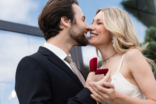 Smiling man in suit holding engagement ring and kissing girlfriend on terrace.
