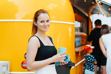 Smiling red-haired young woman holding cup of drink while standing near yellow food truck in park and looking at camera. Cute female customer in line for food outdoors