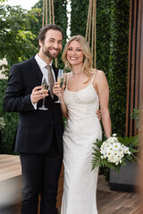 Positive newlyweds with champagne and bouquet looking at camera on terrace.