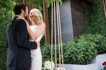 Smiling newlyweds embracing near bouquet on swing on terrace.