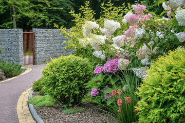 Close up fragment of the garden with lot of decorative green plants, bushes and flowers, and paved lane leading to the house entrance gates. Selective focus.