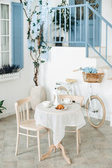 Romantic seating area with beige chairs and a round vintage table, on a blurred background of a white house exterior with a blue door and window and a bicycle with flowers in a basket at the wall