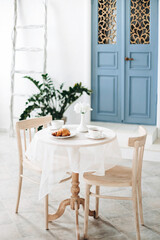 Fototapeta na wymiar Romantic seating area with beige chairs and a round vintage table, against a blurred background of a white mediterranean house with a blue door and a potted plant near the wall. Patio of Santorini