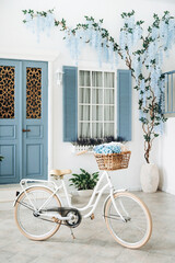 Fototapeta na wymiar Vintage white bike with flowers in a basket against the blured background of a white mediterranean house with a blue door and window and flowering tree on the wall.