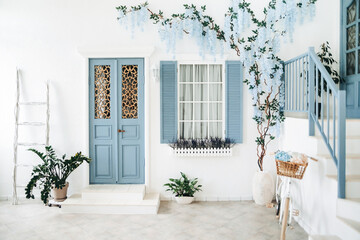 Exterior of a white mediterranean-style house with a blue door and a window, a flowering tree. Traditional patio of Santorini - Powered by Adobe