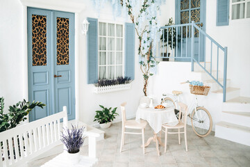 Mediterranean style white house exterior with blue door and window, flowering tree and romantic seating area. Traditional patio of Santorini