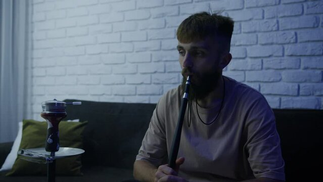 A bearded stylish man is sitting on the couch at home and smoking a hookah