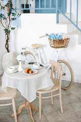 Romantic seating area with beige chairs and round vintage table with two cups of coffee and croissant, on blurred background of a white bicycle with flowers in a basket leaning against a white wall