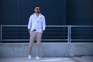 Cool modern young hipster man with a stylish hairstyle in sunglasses in a white shirt. Attractive urban guy posing near a dark wall. Fashionable spring menswear.