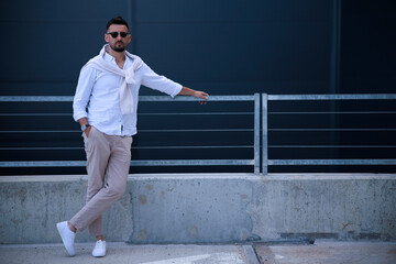 Fototapeta na wymiar Cool modern young hipster man with a stylish hairstyle in sunglasses in a white shirt. Attractive urban guy posing near a dark wall. Fashionable spring menswear.