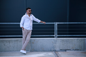 Young stylish brutal men in stylish clothes in sunglasses stands is in a city near a gray building. American guy. Fashionable men's summer clothes. Youth style.