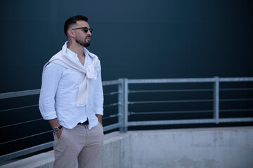 Young stylish brutal men in stylish clothes in sunglasses stands is in a city near a gray building. American guy. Fashionable men's summer clothes. Youth style.