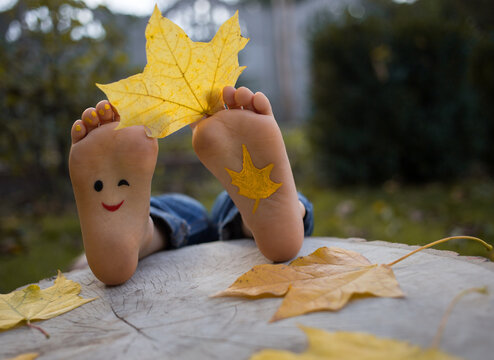 yellow autumn leaf between the toes on the bare feet of a toddler child. painted smile on the feet. indulge, positive thinking, happy childhood. Hello, Autumn. seasonal fun photo ideas