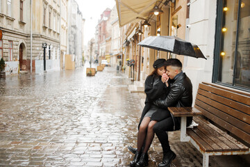 Fototapeta na wymiar Pretty couple sitting near a cafe on a bench under an umbrella in rainy weather. Concept of love, romance and passion.
