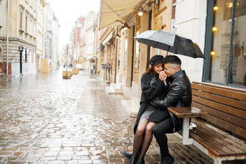 Fototapeta na wymiar Happy couple sitting near a cafe on a bench under an umbrella in rainy weather. Concept of love, romance and passion.