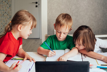 Children sit at a table with pens and notebooks and use a laptop. Homeschooling concept, learning online