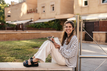Happy young caucasian girl draws on digital tablet with stylus while sitting outdoors. Brown-haired...