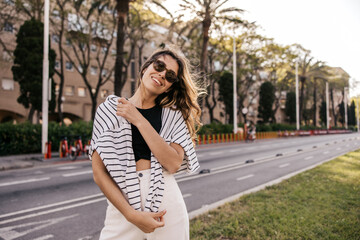 Beautiful young caucasian girl in sunglasses posing at camera walking outdoors. Blonde wears top, sweatshirt and pants spring. Relaxation concept