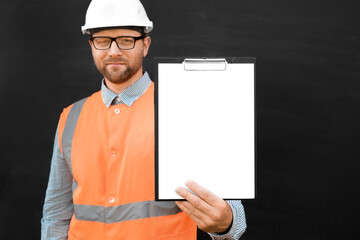 Happy Engineer Pointing At Blank Paper Sheet, Clipboard. Man Builder In Safety Vest And Hard Hat Advertising Empty White Tablet Standing Over Black Background, Mockup. All For Construction, Copy Space