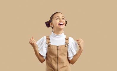 Joyful excited preteen girl clenching fists sincerely rejoices and laughs loudly on beige...