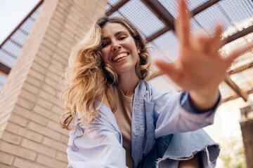 Bottom view of pretty young caucasian girl reaching for camera and smiling relaxing outdoors. Blonde with piercing wears casual shirt. Concept of enjoying moment