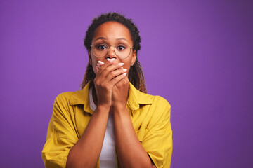 Fototapeta na wymiar Frightened young ethnic African American woman covers mouth with hands experiencing negative emotions restrains himself so as not to scream and wants to avoid panic stands on purple background