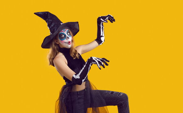 Portrait of funny child in Halloween disguise dancing isolated on yellow color background. Happy little girl in skeleton costume and witch hat, with skull makeup having fun at Halloween party