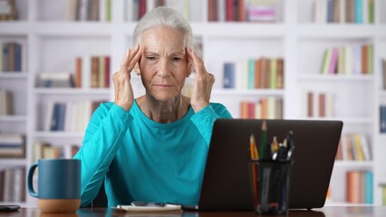 Middle age senior older woman tired with headache online concept, watching business training class, live webinar on laptop computer remote working, social distance learning, writing notes home office.