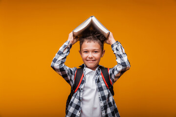 Portrait of a schoolboy boy holding a book over his head, with a bag, backpack, isolated on a yellow background.back to school