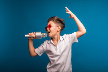 a boy sings into a microphone on a blue background. A handsome boy in a white T-shirt and shorts stands on a blue background and emotionally sings into the microphone. © inna717